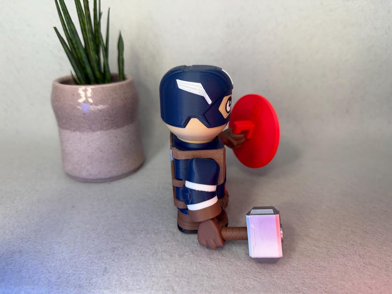 Op de afbeelding: Een Captain America-beeldje met een blauwe helm en schild, met een hamer. Het beeldje draagt een blauw en bruin uniform. Een kleine plant in een pot staat op de achtergrond.