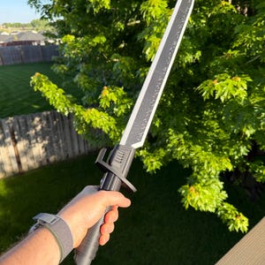 May include: A black and silver sword with a textured blade and handle. The sword has a crossguard and is held in a hand against a background of green foliage and a wooden deck.