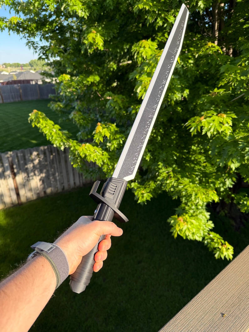 May include: A black and silver sword with a textured blade and handle. The sword has a crossguard and is held in a hand against a background of green foliage and a wooden deck.