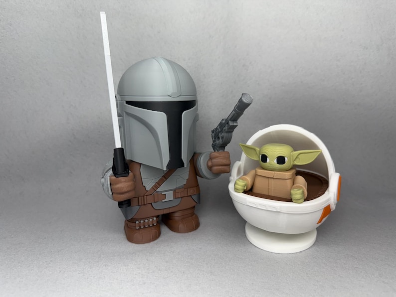 May include: A gray and brown Mandalorian action figure holding a blaster and a white lightsaber. The figure is standing next to a white and orange baby Yoda action figure sitting in a floating pod.
