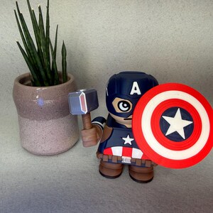 Op de afbeelding: Een Captain America-beeldje met schild en hamer, draagt een blauwe helm met een witte "A". Het beeldje heeft een rood, wit en blauw schild met een witte ster. Links staat een kleine potplant.