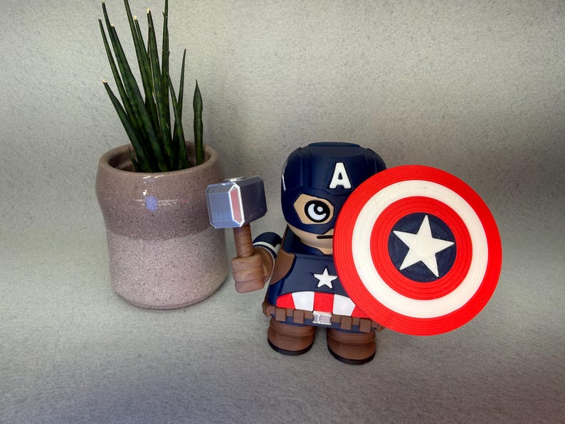 Op de afbeelding: Een Captain America-beeldje met schild en hamer, draagt een blauwe helm met een witte "A". Het beeldje heeft een rood, wit en blauw schild met een witte ster. Links staat een kleine potplant.
