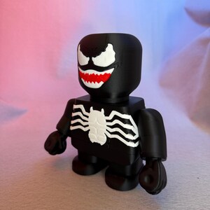 May include: A black and white 3D printed figurine of Venom, a Marvel Comics superhero. The figurine has a large head with a white spider symbol on the chest.
