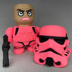 May include: A pink action figure with a black gun, a removable helmet, and a matching pink uniform. The figure has a tan head with an angry expression. The helmet has black accents.