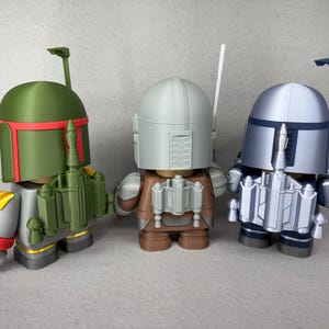 May include: Three Star Wars action figures, Boba Fett, a Mandalorian, and a Stormtrooper, are standing on a gray surface. The figures are made of plastic and are painted in their respective colors. Boba Fett is green, the Mandalorian is brown and gray, and the Stormtrooper is white and gray.