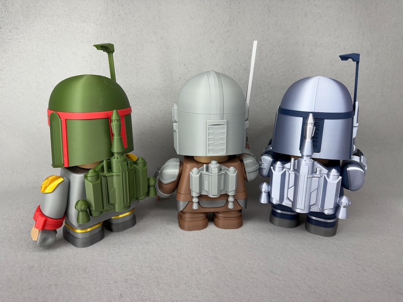 May include: Three Star Wars action figures, Boba Fett, a Mandalorian, and a Stormtrooper, are standing on a gray surface. The figures are made of plastic and are painted in their respective colors. Boba Fett is green, the Mandalorian is brown and gray, and the Stormtrooper is white and gray.