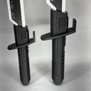 May include: Two black and white toy swords are displayed against a neutral background. The swords have a black handle with a ribbed design, a black guard, and a white blade with a black decorative pattern. The blades are rectangular.