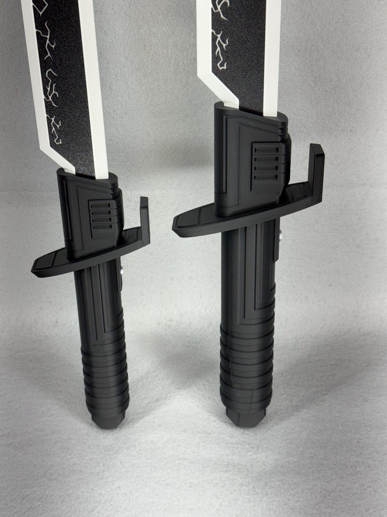 May include: Two black and white toy swords are displayed against a neutral background. The swords have a black handle with a ribbed design, a black guard, and a white blade with a black decorative pattern. The blades are rectangular.