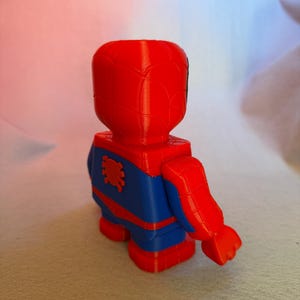 May include: A red and blue 3D printed figurine of Spider-Man. The figurine is posed with one arm raised and the other arm at its side. The figurine has a spider symbol on its chest.