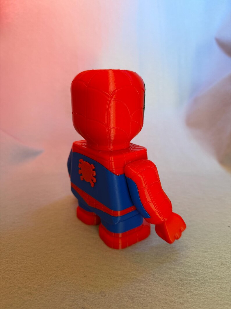 May include: A red and blue 3D printed figurine of Spider-Man. The figurine is posed with one arm raised and the other arm at its side. The figurine has a spider symbol on its chest.