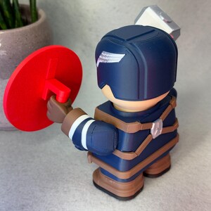 Op de afbeelding: 3D-geprinte beeldje van een superheld in een blauw en bruin uniform, met een rood schild. Het beeldje heeft een wit vleugelontwerp op de helm en een zilveren hamer op de rug.
