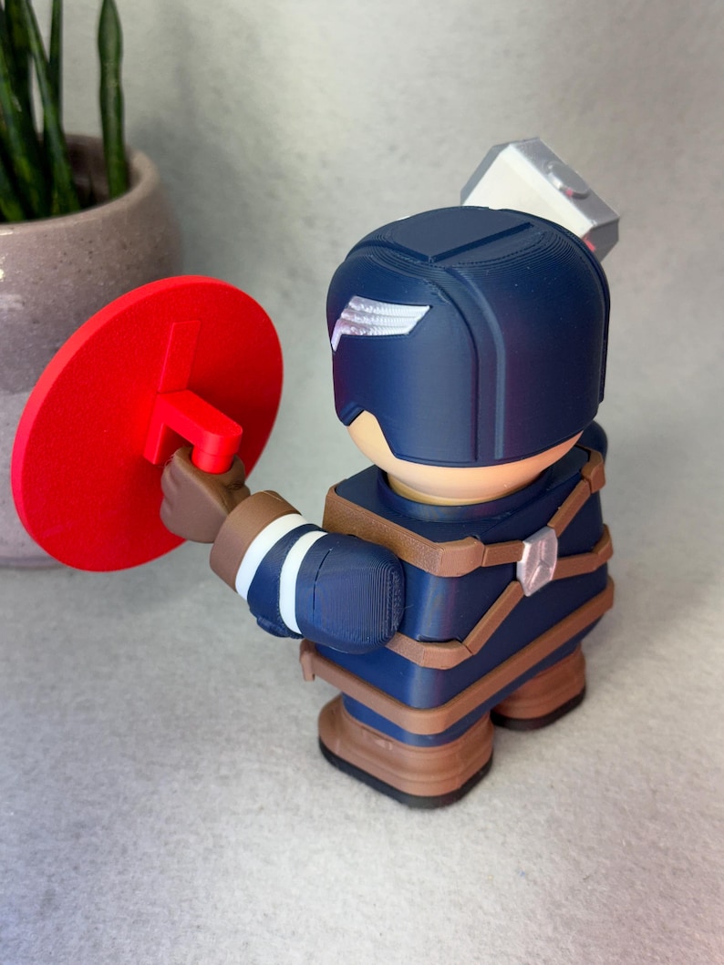 Op de afbeelding: 3D-geprinte beeldje van een superheld in een blauw en bruin uniform, met een rood schild. Het beeldje heeft een wit vleugelontwerp op de helm en een zilveren hamer op de rug.