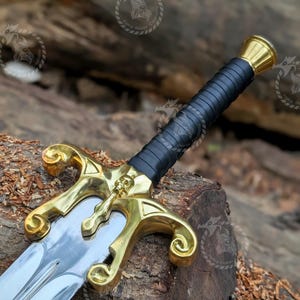 May include: A sword featuring a polished silver-tone blade and a gold-tone metal crossguard with a central skull detail. The handle is wrapped in black material and finished with a gold-tone pommel. The design includes curved quillons.