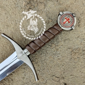 Pode incluir: Uma espada com lâmina de aço polido, punho revestido em couro castanho e guarda de prata. O pomo apresenta um emblema circular com uma cruz vermelha num escudo. Um logótipo circular com um capacete viking e o texto Alpha Norse Creations.