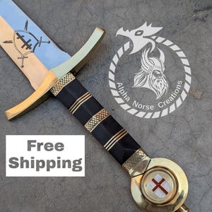 Könnte beinhalten: Ein Schwert im mittelalterlichen Stil mit einem schwarz-goldenen Griff und einem goldenen Knauf mit einem roten Kreuz. Die Klinge ist silberfarben und hat ein Design mit einem Schild mit einem Kreuz darauf. Das Schwert befindet sich auf einem grauen Hintergrund mit dem Text "Kostenloser Versand" und einem Logo für Alpha Norse Creations.