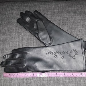 May include: A pair of black gloves with a decorative floral design on the cuff. The gloves are made of a smooth material and are shown with a measuring tape, indicating a length of approximately 10 inches.