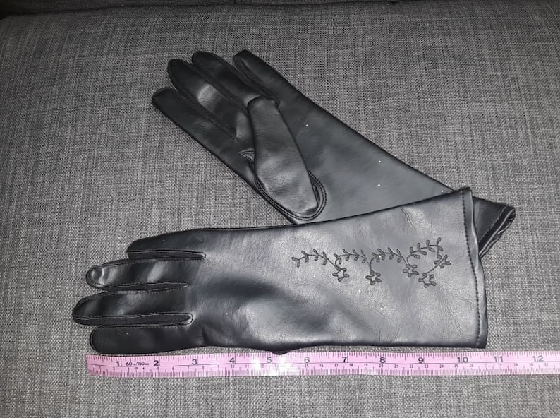 May include: A pair of black gloves with a decorative floral design on the cuff. The gloves are made of a smooth material and are shown with a measuring tape, indicating a length of approximately 10 inches.
