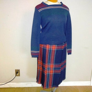 May include: A vintage two-piece outfit featuring a navy blue sweater with red, white, and green stripes on the neckline and cuffs. The outfit includes a pleated plaid skirt in navy blue, red, and orange.
