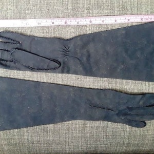 May include: A pair of long, dark blue gloves. The gloves are made of a soft material and have a seam detail along the edges. A pink and white ruler is placed above the gloves.