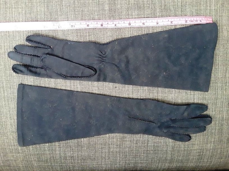 May include: A pair of long, dark blue gloves. The gloves are made of a soft material and have a seam detail along the edges. A pink and white ruler is placed above the gloves.