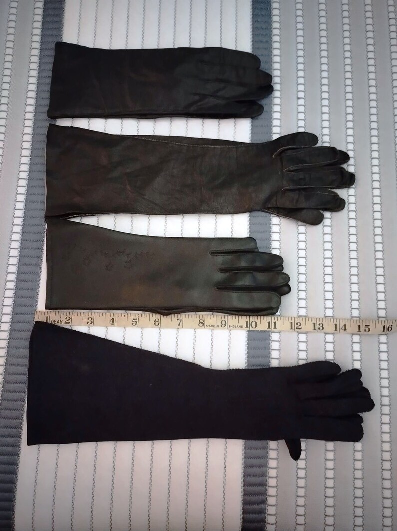 May include: Three pairs of black gloves are displayed. The top two pairs appear to be leather, with the middle pair showing some wear. The bottom pair is a darker, possibly fabric, material. A measuring tape is placed along the gloves.