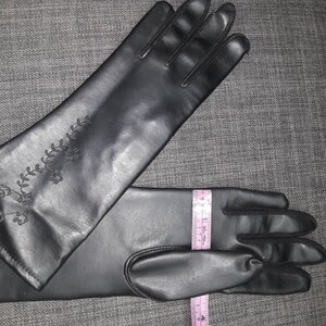 May include: Black gloves with an embroidered floral design on the upper arm. The gloves are measured with a tape measure, approximately 15 inches long.