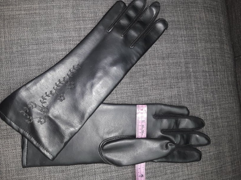 May include: Black gloves with an embroidered floral design on the upper arm. The gloves are measured with a tape measure, approximately 15 inches long.