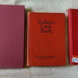 Könnte beinhalten: Drei Kochbücher im Vintage-Stil werden gezeigt. Das erste ist einfarbig mauve, das zweite orange mit dem Text "Watkins Cook Book" und das dritte rot mit einem goldenen Design. Die Bücher sind nebeneinander angeordnet.