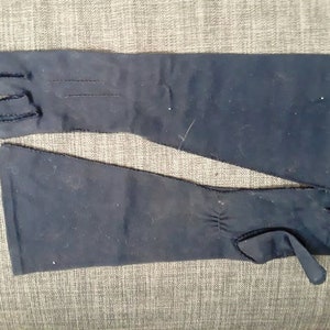 May include: A pair of navy blue, long opera gloves. The gloves are made of a smooth fabric and have a seam detail on the back of the hand. The gloves extend past the wrist and have a small tab detail at the wrist.