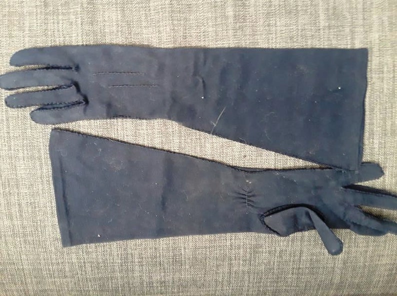 May include: A pair of navy blue, long opera gloves. The gloves are made of a smooth fabric and have a seam detail on the back of the hand. The gloves extend past the wrist and have a small tab detail at the wrist.