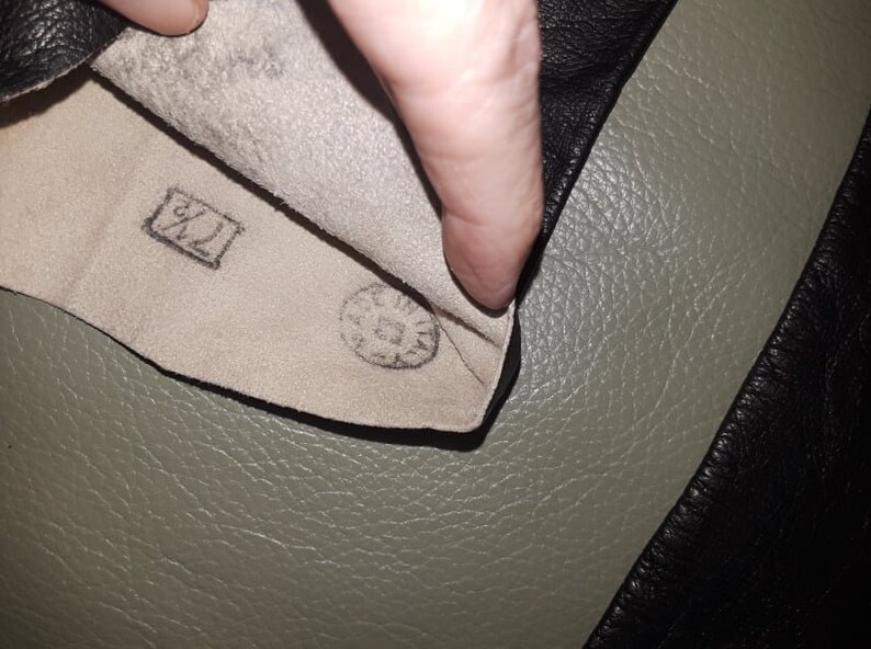 May include: Close-up of the inside of a black leather glove. The interior is a light beige color with a stamped size marking of "7 1/2" in a rectangular box. A circular stamp is also visible.