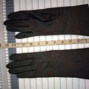 May include: A pair of dark brown gloves, likely leather, with a length of approximately 13 inches. The gloves are laid flat on a striped fabric background, showcasing their sleek design and potential for formal wear or cold weather protection.
