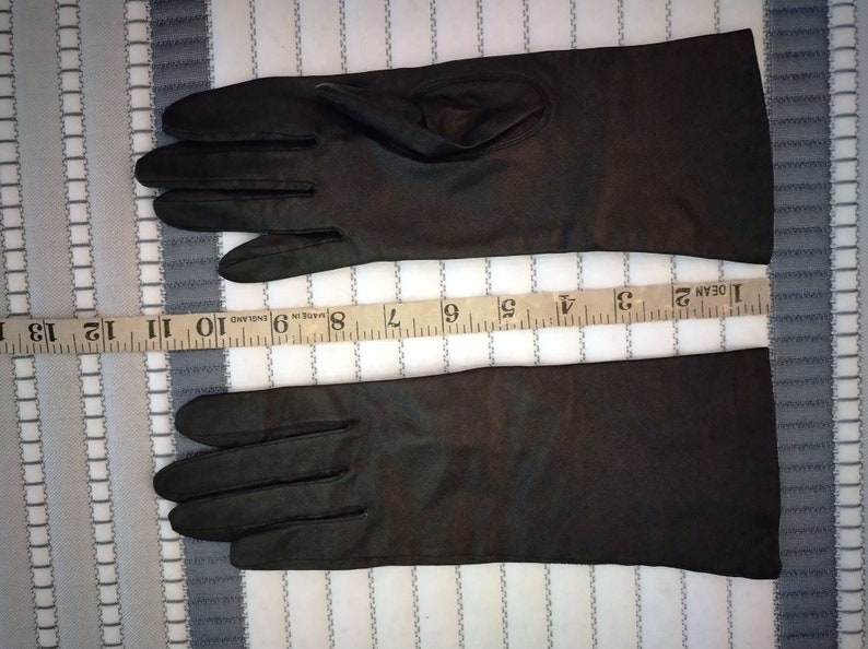 May include: A pair of dark brown gloves, likely leather, with a length of approximately 13 inches. The gloves are laid flat on a striped fabric background, showcasing their sleek design and potential for formal wear or cold weather protection.