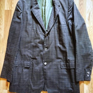 May include: A brown and black plaid blazer with a notched collar, two front pockets, and button closures. The blazer has a green lining and three buttons on each cuff. The jacket is laid flat on a wooden surface.