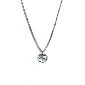 May include: A silver necklace featuring a textured, circular pendant. The pendant is suspended from a fine chain, offering a minimalist aesthetic. The necklace is presented on a plain white backdrop.
