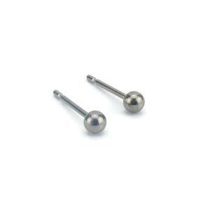 Può includere: Un paio di piccoli orecchini a lobo color argento. Ogni orecchino presenta una sfera rotonda e lucida all'estremità di un perno sottile e dritto. Gli orecchini sono esposti su sfondo bianco.
