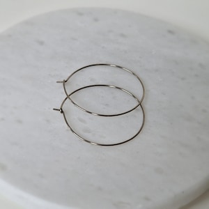 Peut inclure: Deux boucles d'oreilles créoles argentées. Les boucles d'oreilles sont circulaires et semblent être faites d'un fil métallique fin. Elles sont présentées sur une surface en marbre blanc.