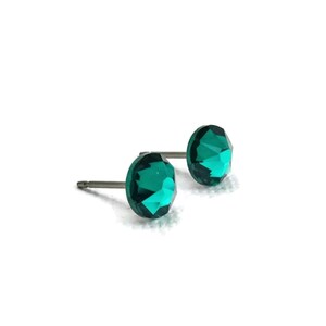 May include: A pair of emerald green stud earrings. Each earring features a faceted, round gemstone set on a silver-colored post. The earrings are displayed against a white background.