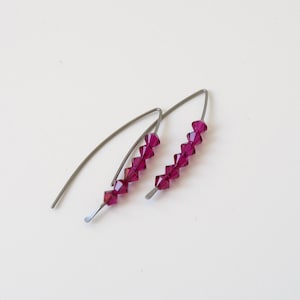 May include: A pair of dangle earrings featuring a silver-coloured metal hook and a row of faceted, deep magenta beads. The earrings are presented against a white backdrop.