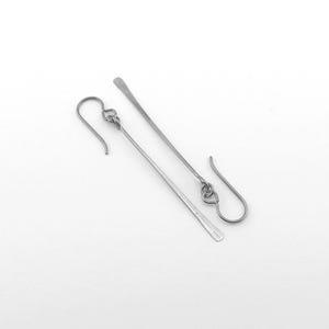 May include: A pair of silver-toned dangle earrings. Each earring features a long, slender bar suspended from a decorative loop and an ear wire. The earrings are displayed against a white background, highlighting their minimalist design.