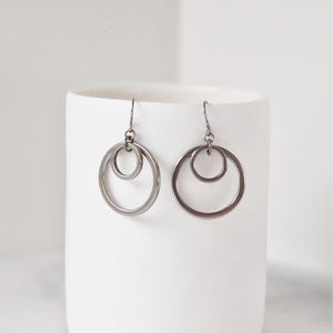 May include: A pair of silver-toned earrings with a double-circle design. Each earring features a large, open circle with a smaller circle nested inside. The earrings hang from simple hooks, showcasing a minimalist and modern aesthetic.