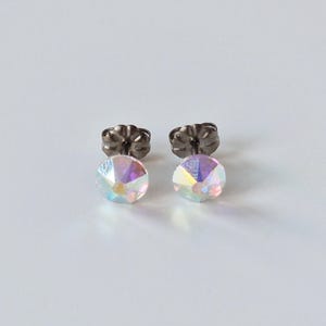 May include: A pair of iridescent crystal stud earrings with dark gray backs. The round crystals reflect multiple colors, creating a shimmering effect. The earrings are set against a white background.