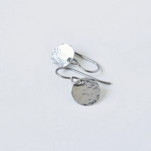 May include: A pair of silver-colored disc earrings with a hammered texture. The earrings have a simple, minimalist design with a small, round disc hanging from a curved ear wire. The earrings are displayed on a white surface.