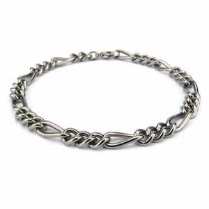 Op de afbeelding: Een zilverkleurige kettingarmband met een Figaro-schakels ontwerp. De armband heeft een patroon van drie kleine cirkelvormige schakels, gevolgd door één langwerpige ovale schakel. Het metaal heeft een gepolijste afwerking, die licht reflecteert.
