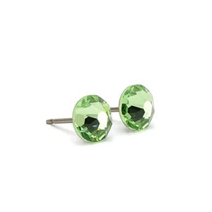 May include: A pair of light green stud earrings. Each earring features a faceted, round, light green crystal. The earrings are set on silver-colored posts. The background is plain white.