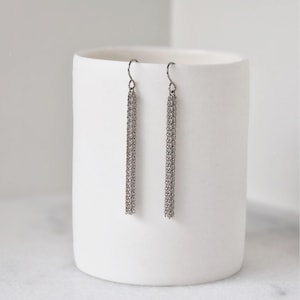 May include: A pair of silver-toned chain tassel earrings. The earrings hang from a simple hook and feature multiple strands of delicate chains. The earrings are displayed against a white background.