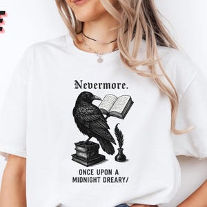 May include: White t-shirt featuring a black illustration of a raven perched on books, holding an open book. The text "Nevermore" and "Once Upon a Midnight Dreary" are also printed on the shirt. The word "WHITE" is printed in the upper left corner.
