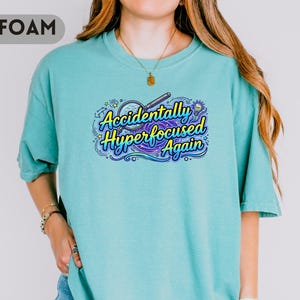 May include: A seafoam green short-sleeve t-shirt featuring a graphic print of a magnifying glass over a swirling purple and blue galaxy design. The text reads "Accidentally Hyperfocused Again" in a stylized yellow and blue font with small stars.