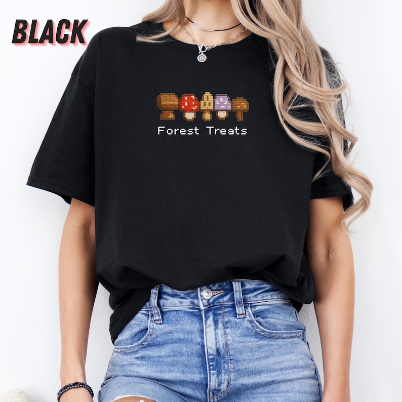 May include: Black t-shirt featuring a pixelated mushroom graphic in brown, red, yellow, and purple. The text "Forest Treats" is printed below the graphic. The shirt is a casual style.