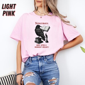 May include: Light pink t-shirt featuring a black illustration of a raven perched on books, holding a book and quill. The text "Nevermore" and "Once Upon a Midnight Dreary" are printed above and below the raven design. The words "Light Pink" are printed in the upper left corner.